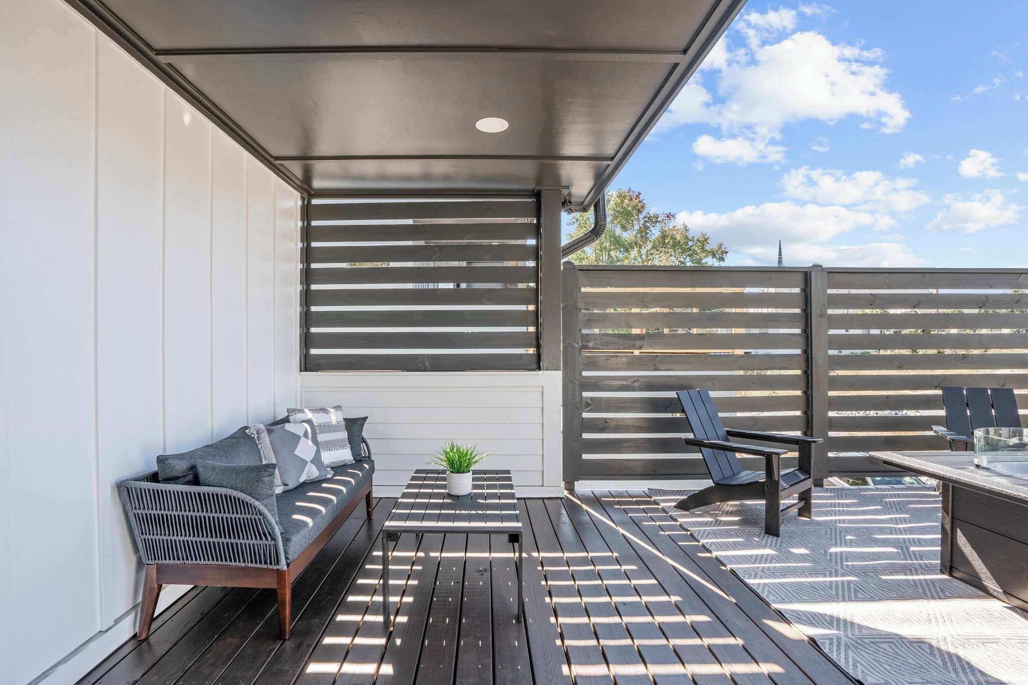 An outdoor patio with a wooden deck, a sofa, a small table with a plant, and two chairs.