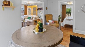 En rummelig stue med et rundt spisebord, en sofa og en lænestol. Et vindue med udsigt over grønne omgivelser.