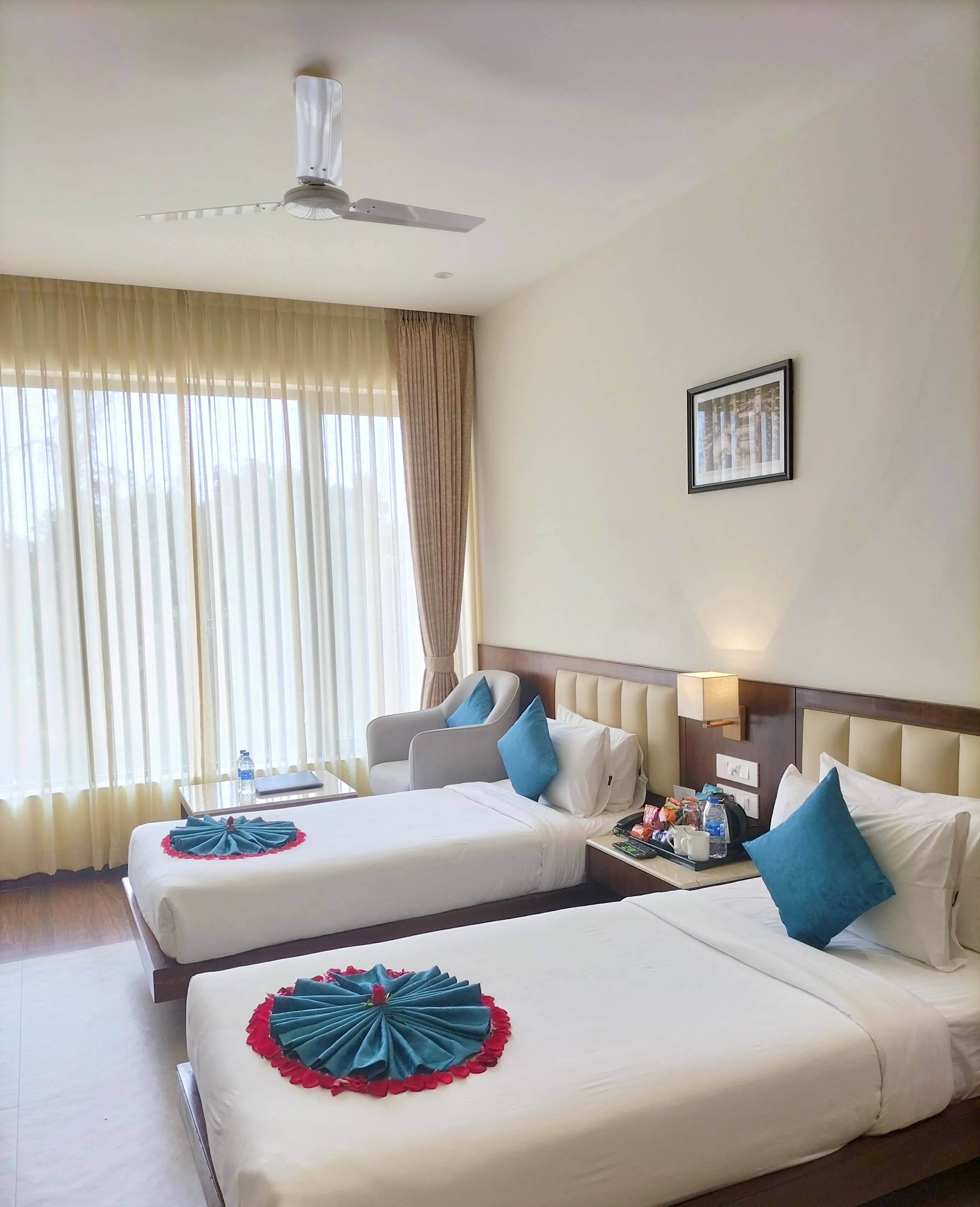 A hotel room with two beds, a ceiling fan, a framed picture on the wall, and a nightstand with a lamp and a tray of snacks.