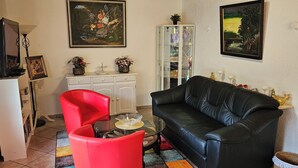 Ein Wohnzimmer mit einer schwarzen Ledersofa, einem roten Sessel, einem Glastisch und einem weißen Schrank. An der Wand hängen zwei Bilder und auf dem Schrank steht eine kleine Pflanze.