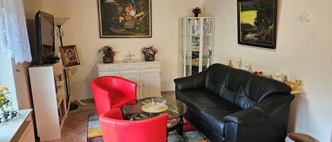 A living room with a black leather sofa, a red chair, a glass coffee table, and a white cabinet. There are two paintings on the wall and a small plant on the cabinet.