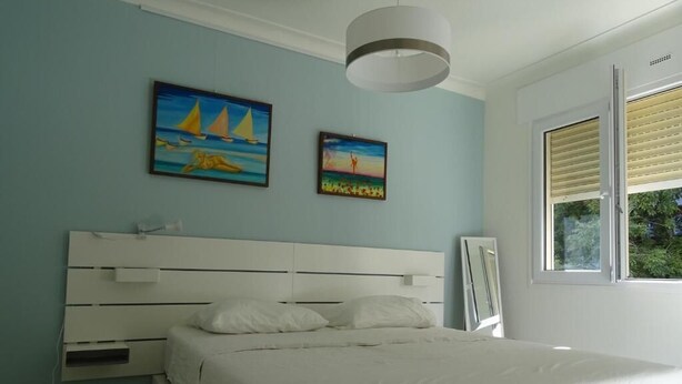 A bedroom with a white bed, a nightstand, a painting of sailboats, a ceiling light, and a window with blinds. - Enchanting apartment with AC in Cannes (Cannes)