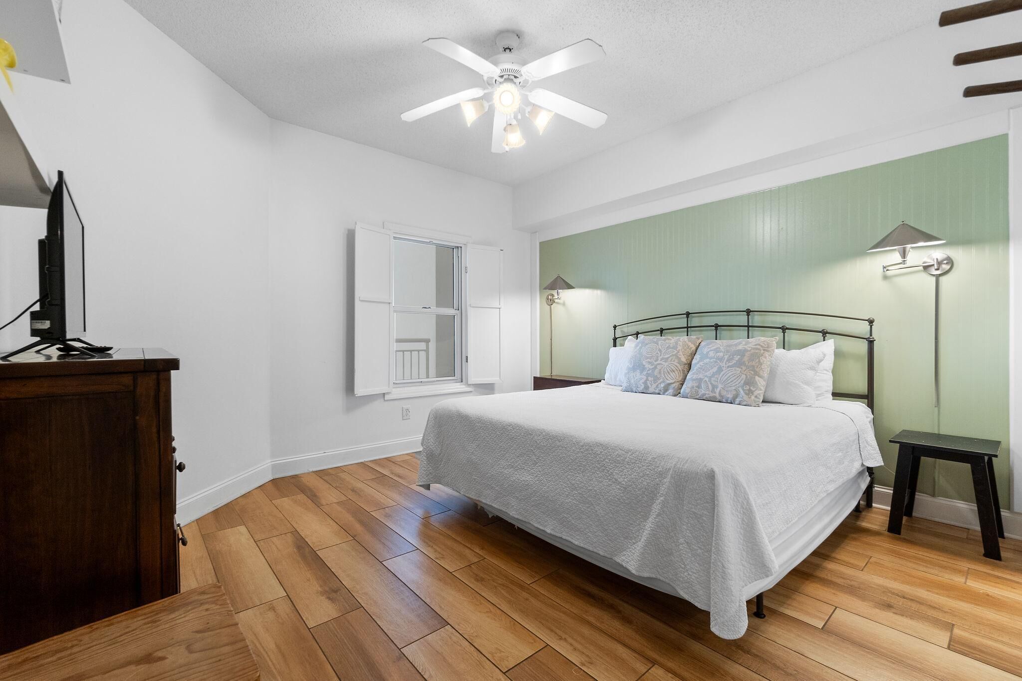 A bedroom with a bed, a ceiling fan, a nightstand, a lamp, and a wooden floor.
