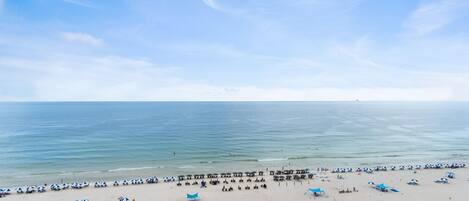 A beach with rows of umbrellas and lounge chairs, a pier extending into the water, and a clear sky.