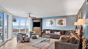 Ein geräumiges Wohnzimmer mit großem Fenster zum Strand, Deckenventilator und Flachbildfernseher.