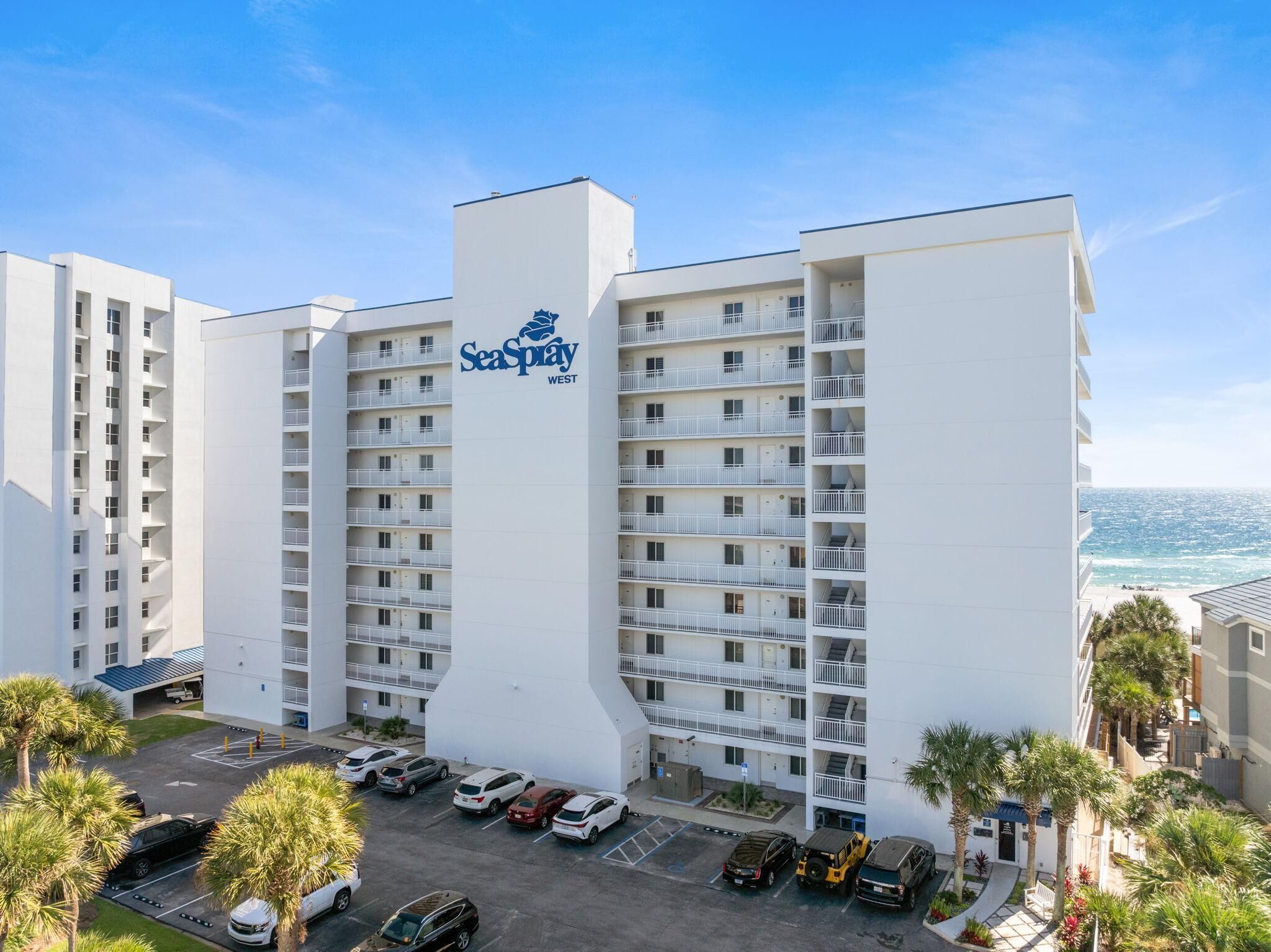 Un hotel de varios pisos con un llamativo letrero SeaSpray WEST, con balcones y un estacionamiento con varios automóviles.
