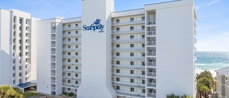 Un hotel de varios pisos con un llamativo letrero SeaSpray WEST, con balcones y un estacionamiento con varios automóviles.