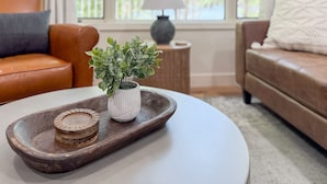 Uma sala de estar com um sofá de couro marrom, uma poltrona branca, uma mesinha pequena com uma planta e uma bandeja de madeira com objetos decorativos.