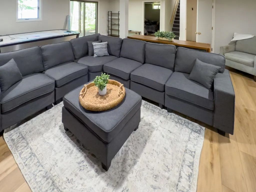 A modern living room with a grey sectional sofa, a small ottoman with a plant, and a white patterned rug.