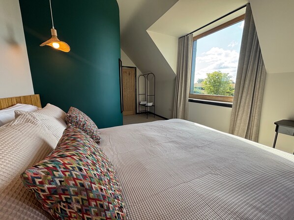 Un dormitorio con cama, una lámpara colgante, un ventanal con cortinas y vistas a la vegetación exterior.