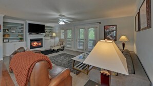 A living room with a fireplace, a television, a ceiling fan, and a large window with blinds. - Riverside Trout Cottage / Sleeps 8 / Short walk to Downtown Helen (Helen)