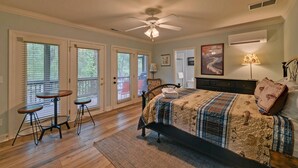 A bedroom with a bed, a desk, two chairs, a ceiling fan, and large windows with blinds.