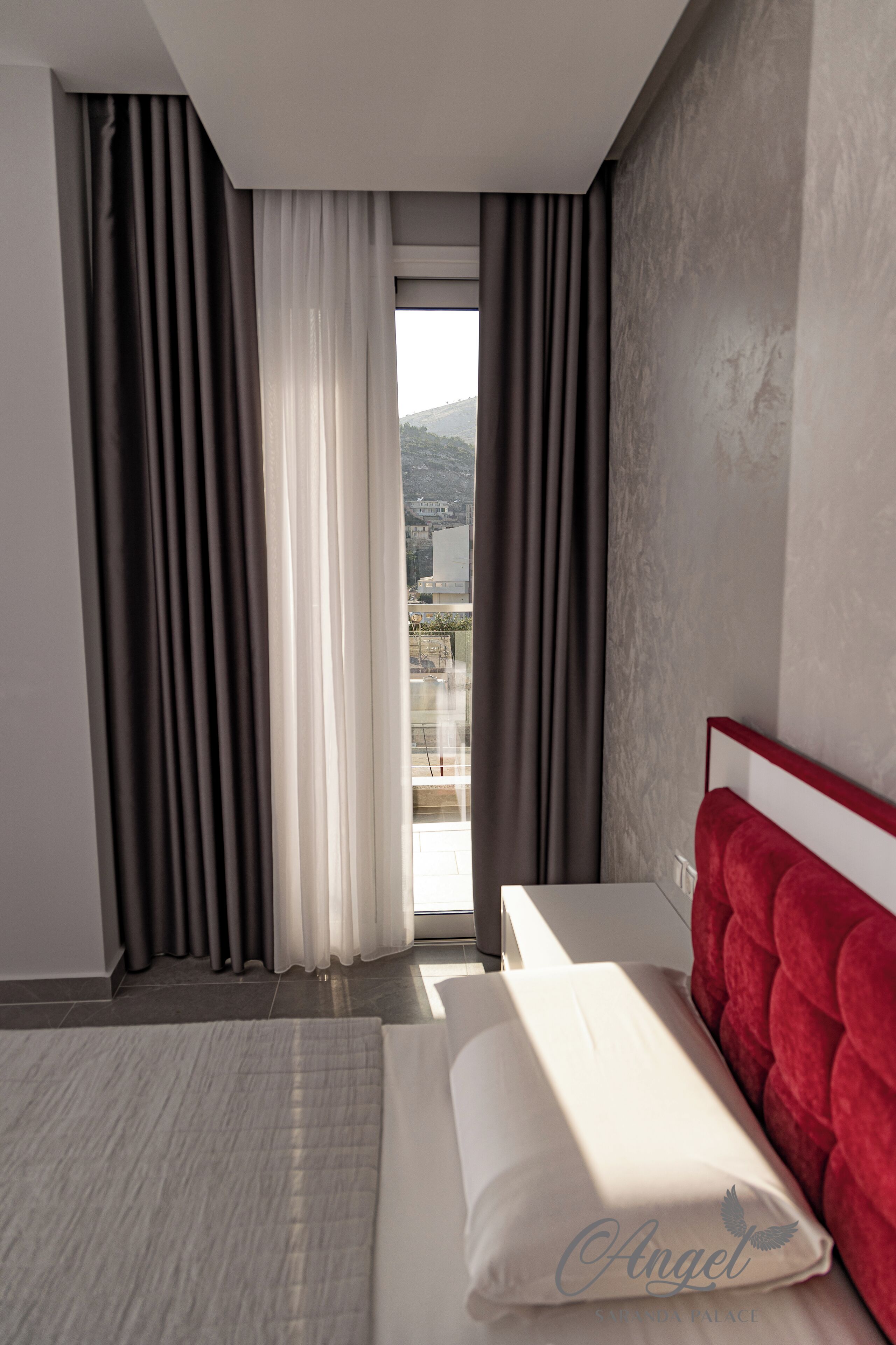 A hotel room with a bed, a red headboard, a window with curtains, and a view of a cityscape.