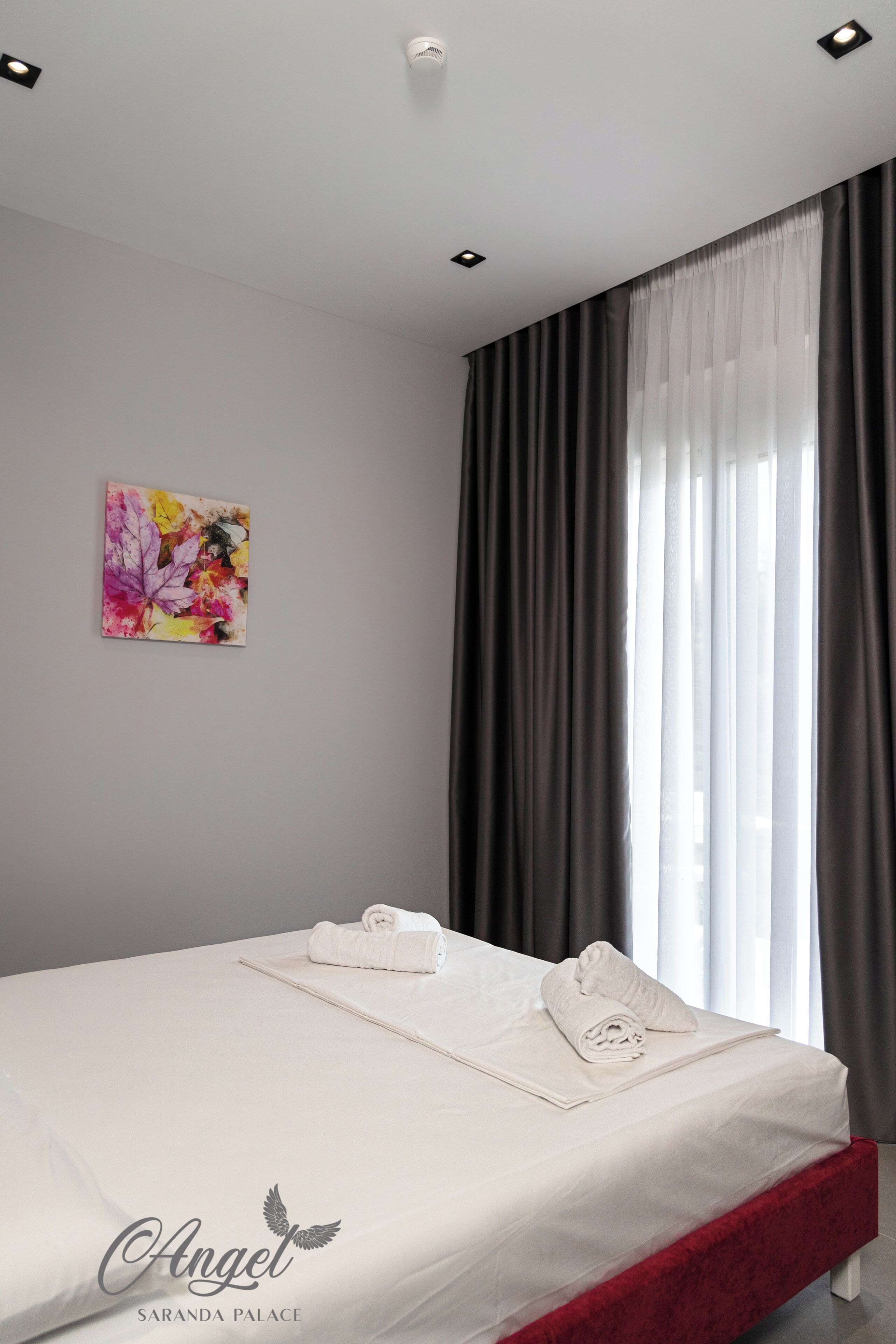 A neatly made bed with white linens and a red headboard, a painting on the wall, and a window with dark curtains.
