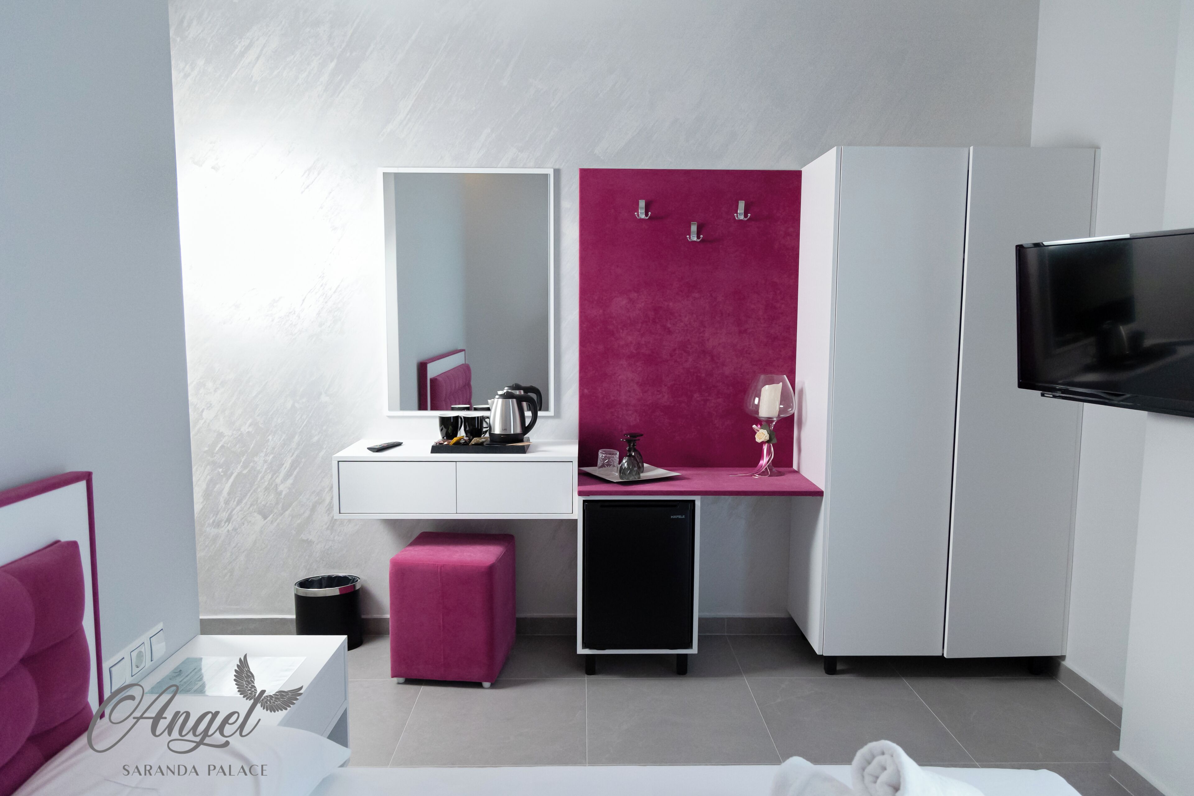 A modern bedroom with a white bed, a pink ottoman, a small black TV, and a white wardrobe.