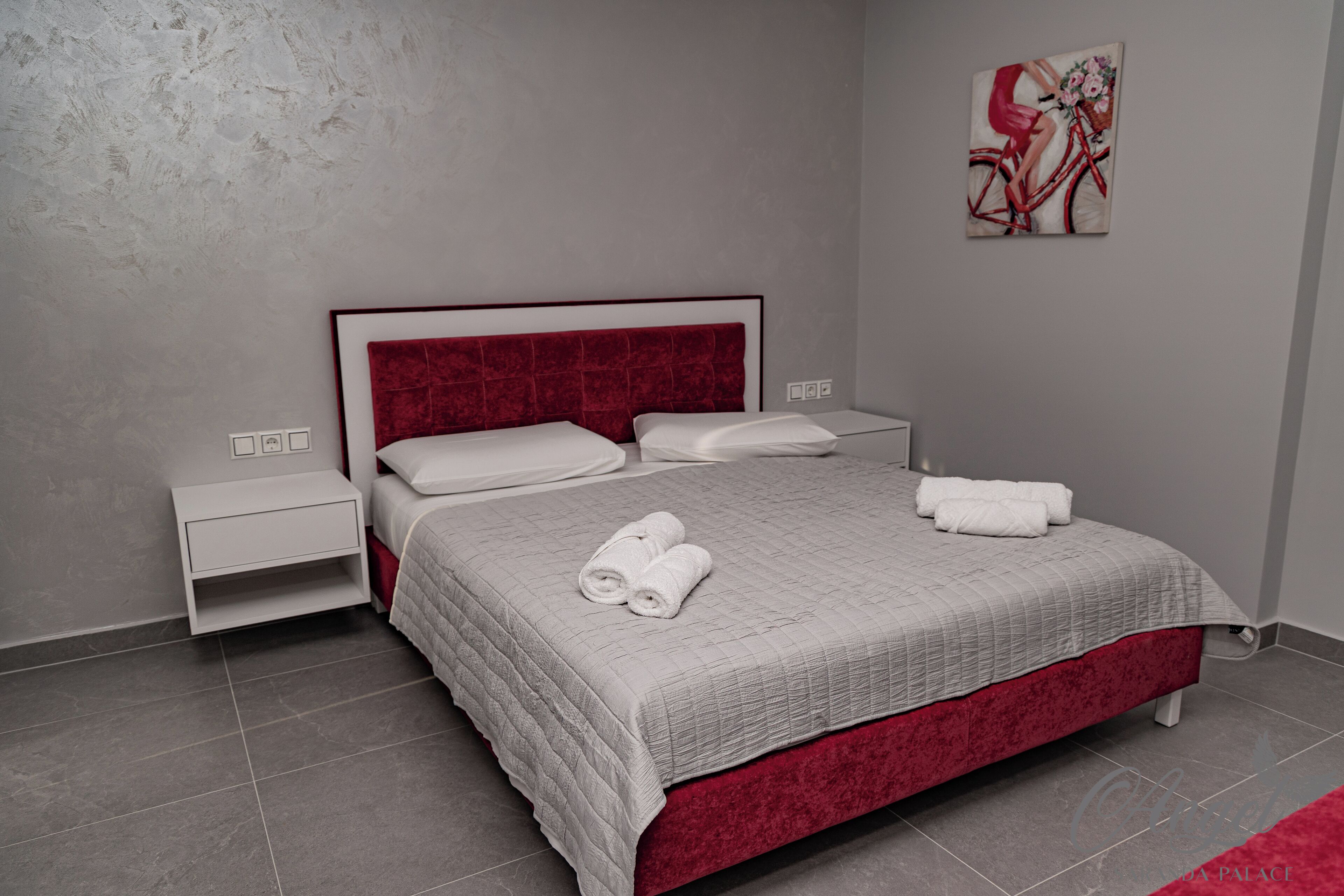 A neatly made bed with a red headboard, white pillows, and a grey quilt. There is a small bedside table with a lamp, and a painting on the wall.