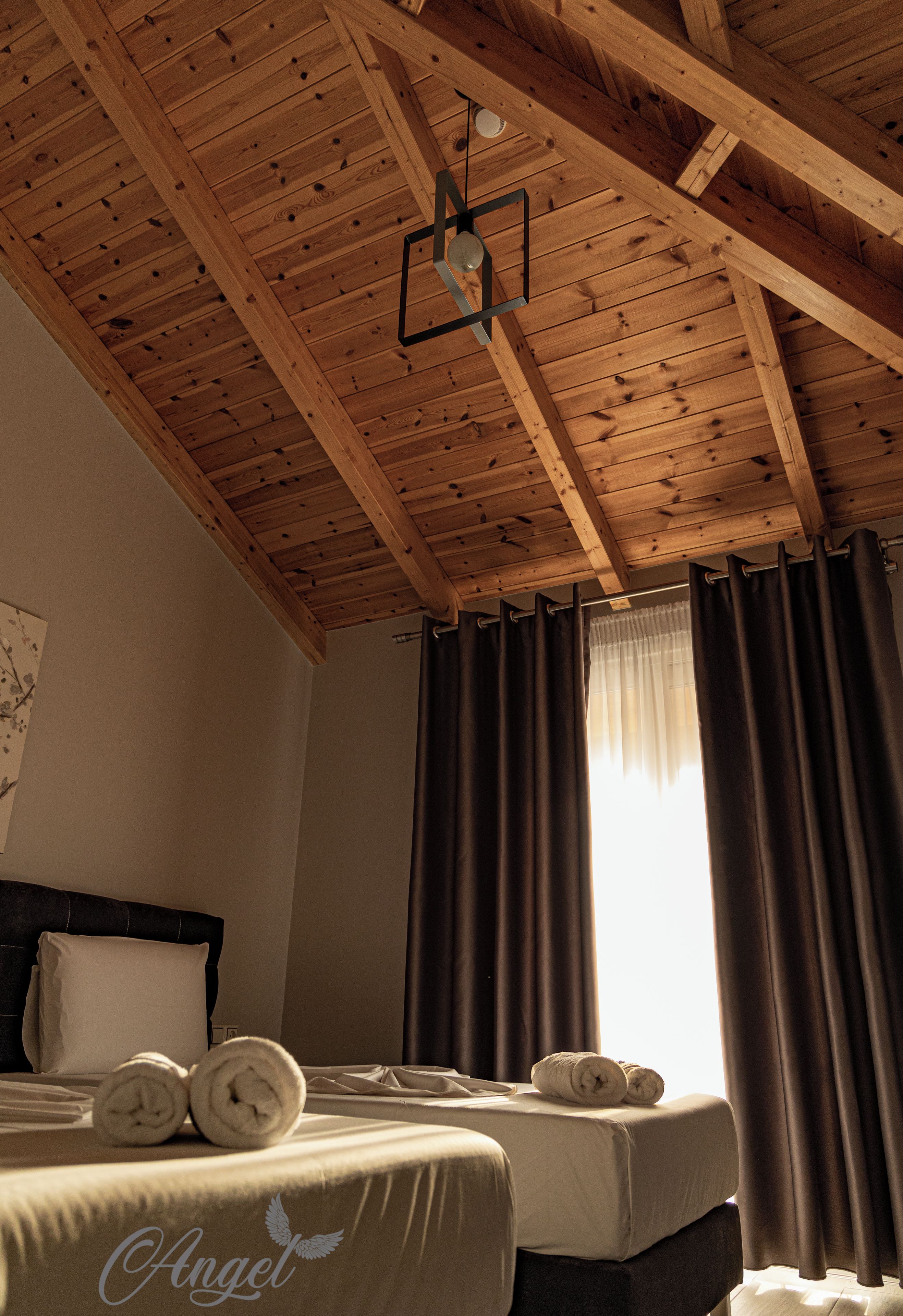 A hotel room with wooden ceiling beams, a bed with rolled towels, and a window with curtains.