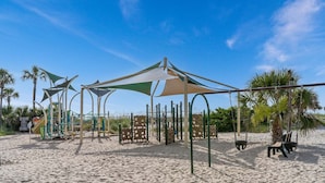 A beach playground with play structures and shaded areas, surrounded by palm trees and sandy ground. - Salty Britches Cottage, with Golf Cart in Ocean Lakes! (Myrtle Beach)
