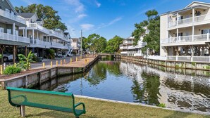 A canal with a bench in the foreground, surrounded by buildings with balconies and a clear sky. - Salty Britches Cottage, with Golf Cart in Ocean Lakes! (Myrtle Beach)