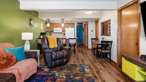 Una sala de estar con sofá, sillón y una alfombra colorida. Se observa una cocina al fondo, con microondas y armarios.