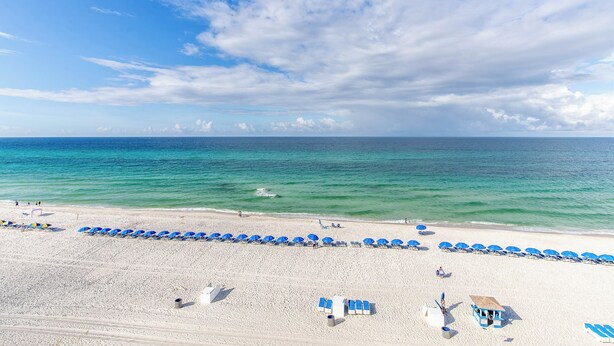 A beach with rows of blue umbrellas and lounge chairs, a lifeguard tower, and a sandy shore. - Coastal Escape West - Unit 506, beach service inc., 1 mile West of Pier Park (Panama City Beach)