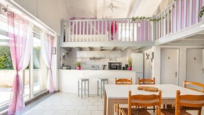Un comedor con mesa blanca y sillas de madera, una cocina con armarios blancos, y un balcón con barandillas moradas.