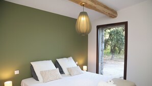 Un dormitorio con cama, almohadas, lámpara de noche y ventana con vistas a árboles.