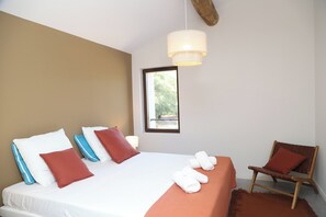 A neatly made bed with white and blue pillows, a wooden chair, and a window with a view of greenery.