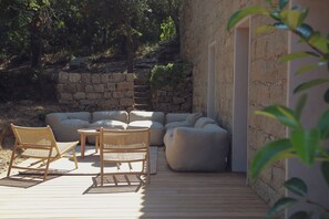 A wooden deck with outdoor furniture, including chairs and a sofa, near a stone wall and a building.
