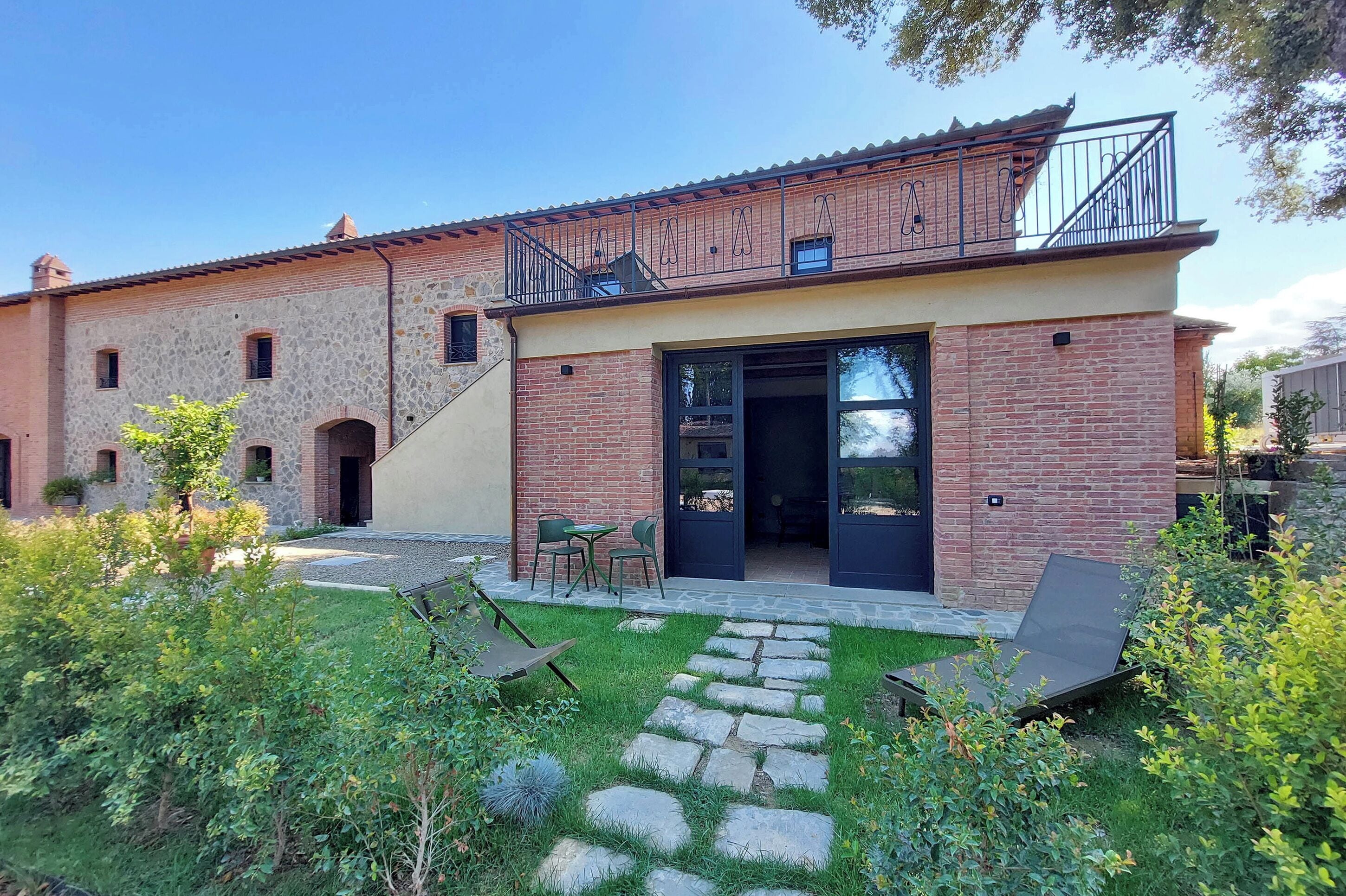A two-story building with a stone facade, a balcony with metal railings, and a paved patio area with outdoor seating.