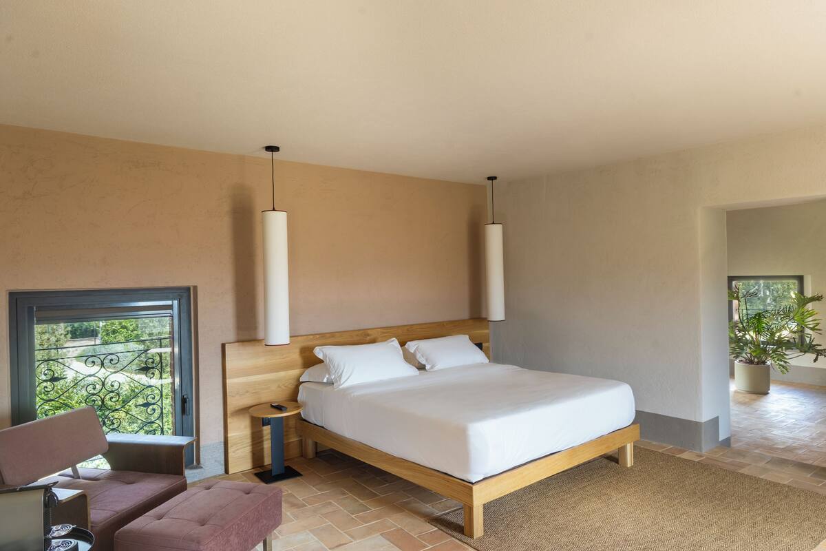 Une chambre à coucher avec un lit en bois, des tables de chevet, un banc, une plante en pot et une vue sur la verdure par la fenêtre.
