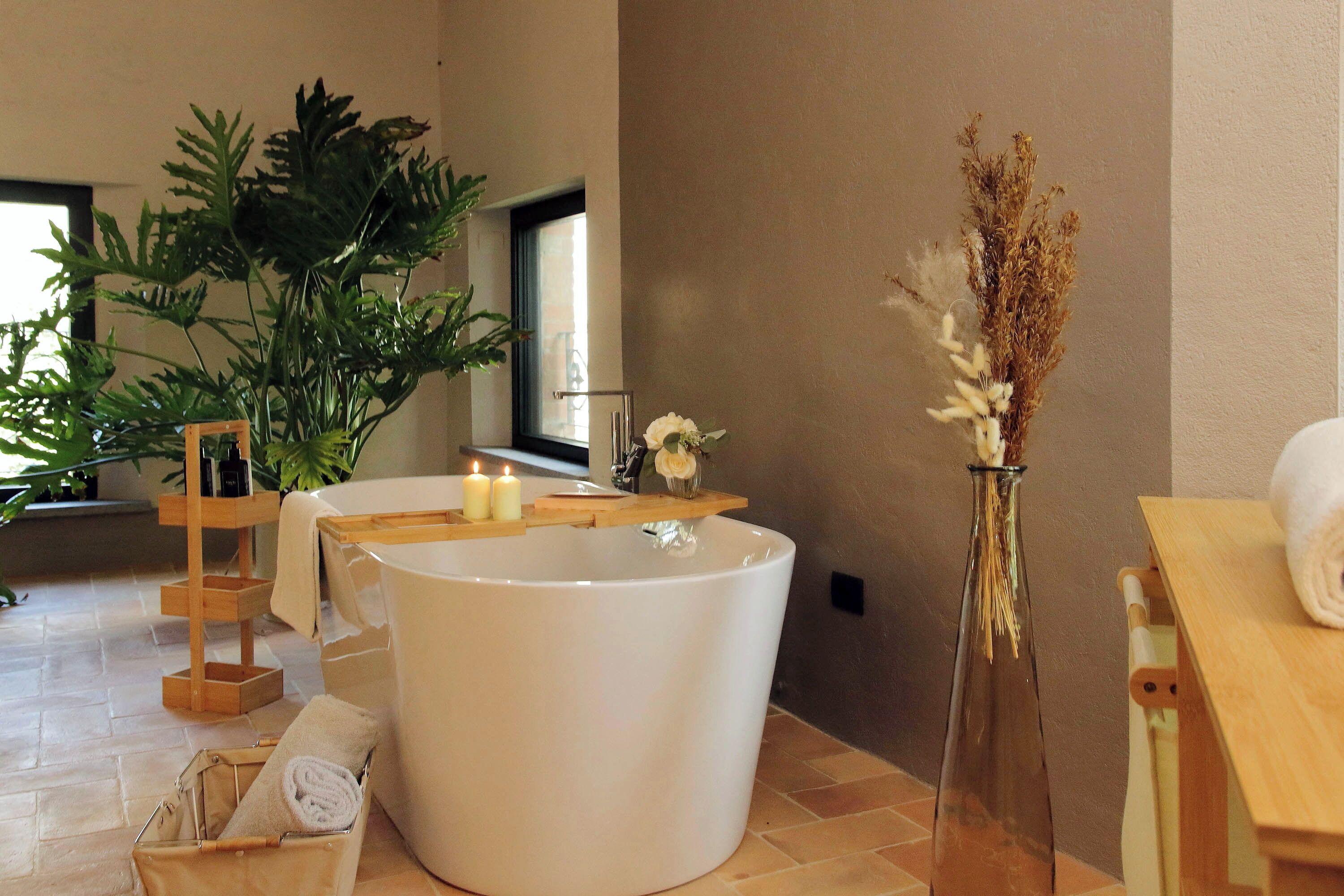 A spa room with a large bathtub, a wooden shelf with candles, a vase with dried flowers, and a wooden bench.