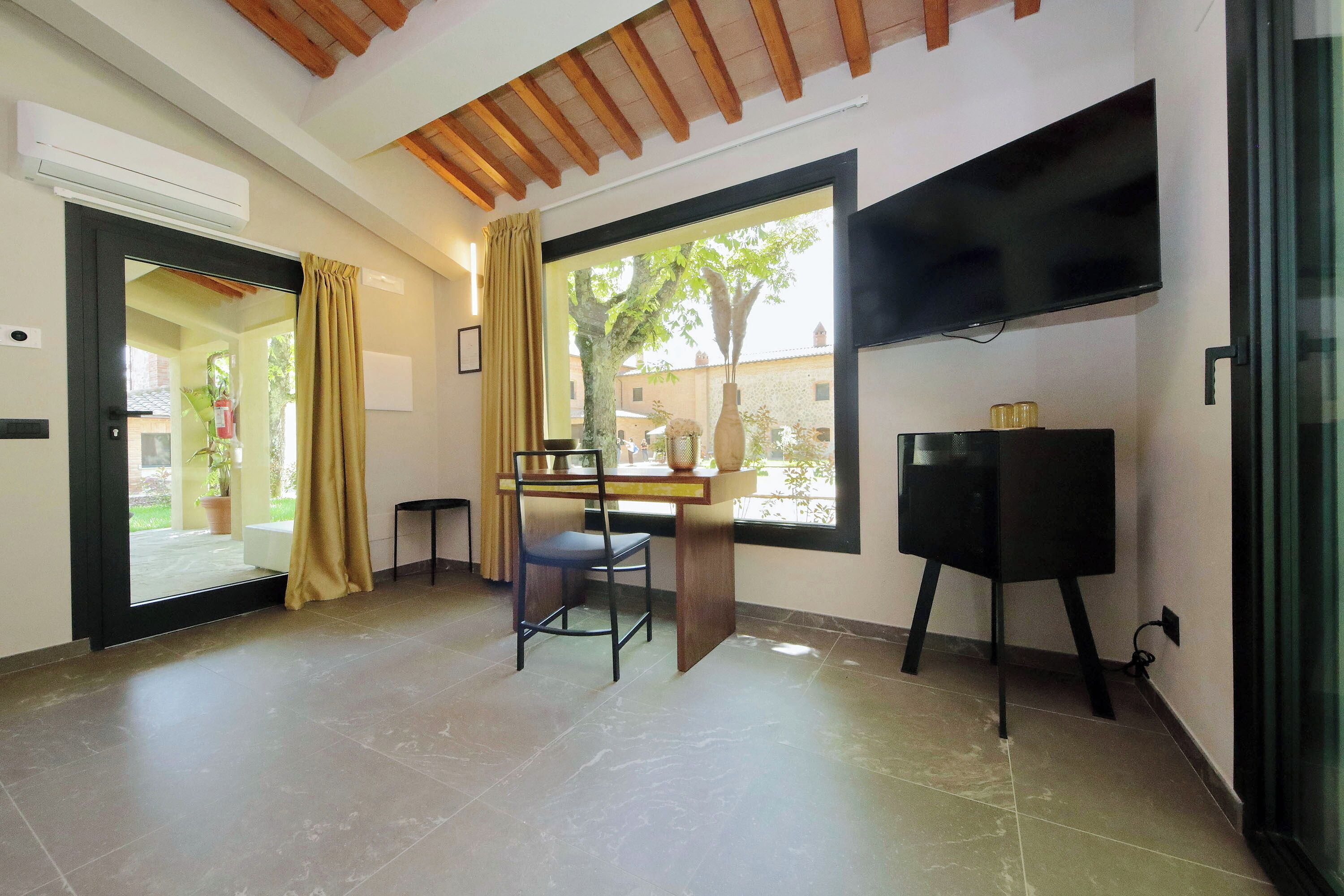 A room with a desk, chair, TV, and a window with a view of greenery.