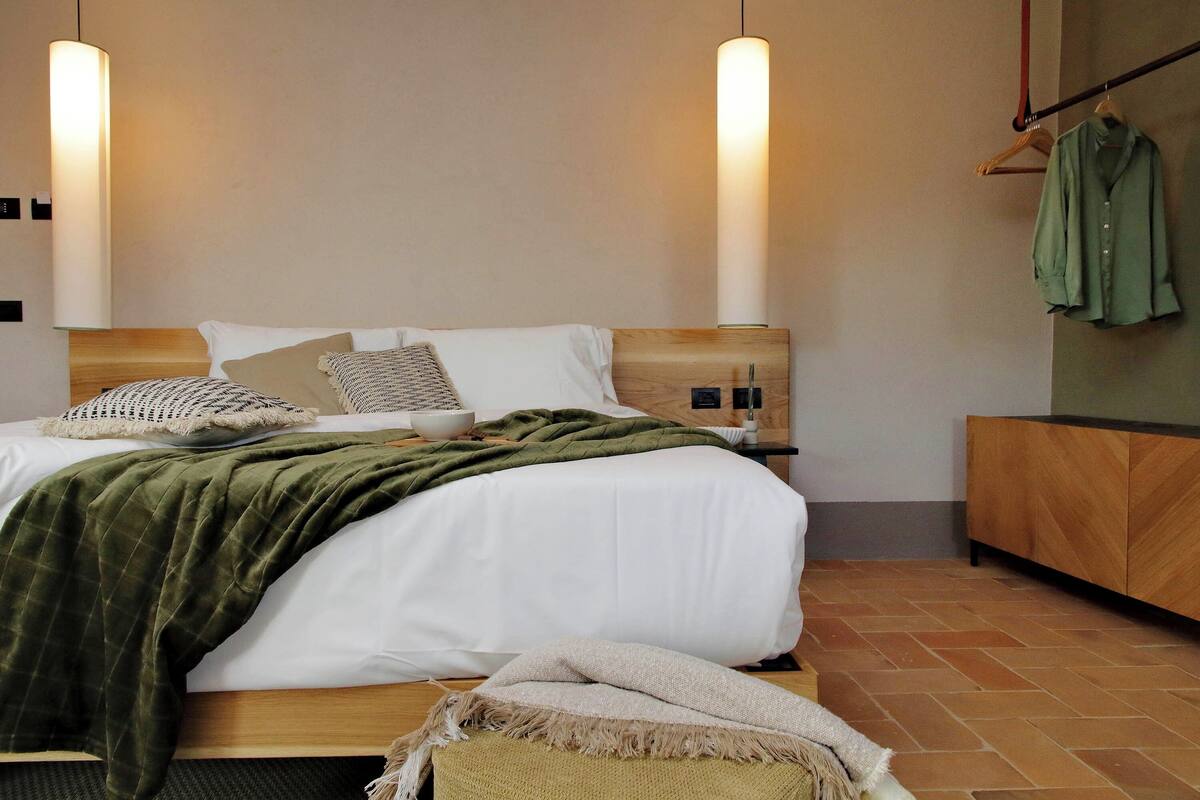 Un lit bien fait, recouvert d’une couverture verte, avec deux lampes de chevet et une table de nuit en bois sur laquelle est posé un porte-vêtements avec une chemise verte.
