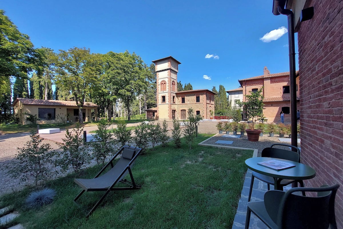 Une cour avec une pelouse, un fauteuil de jardin, une table et un bâtiment en briques surmonté d’une tour.