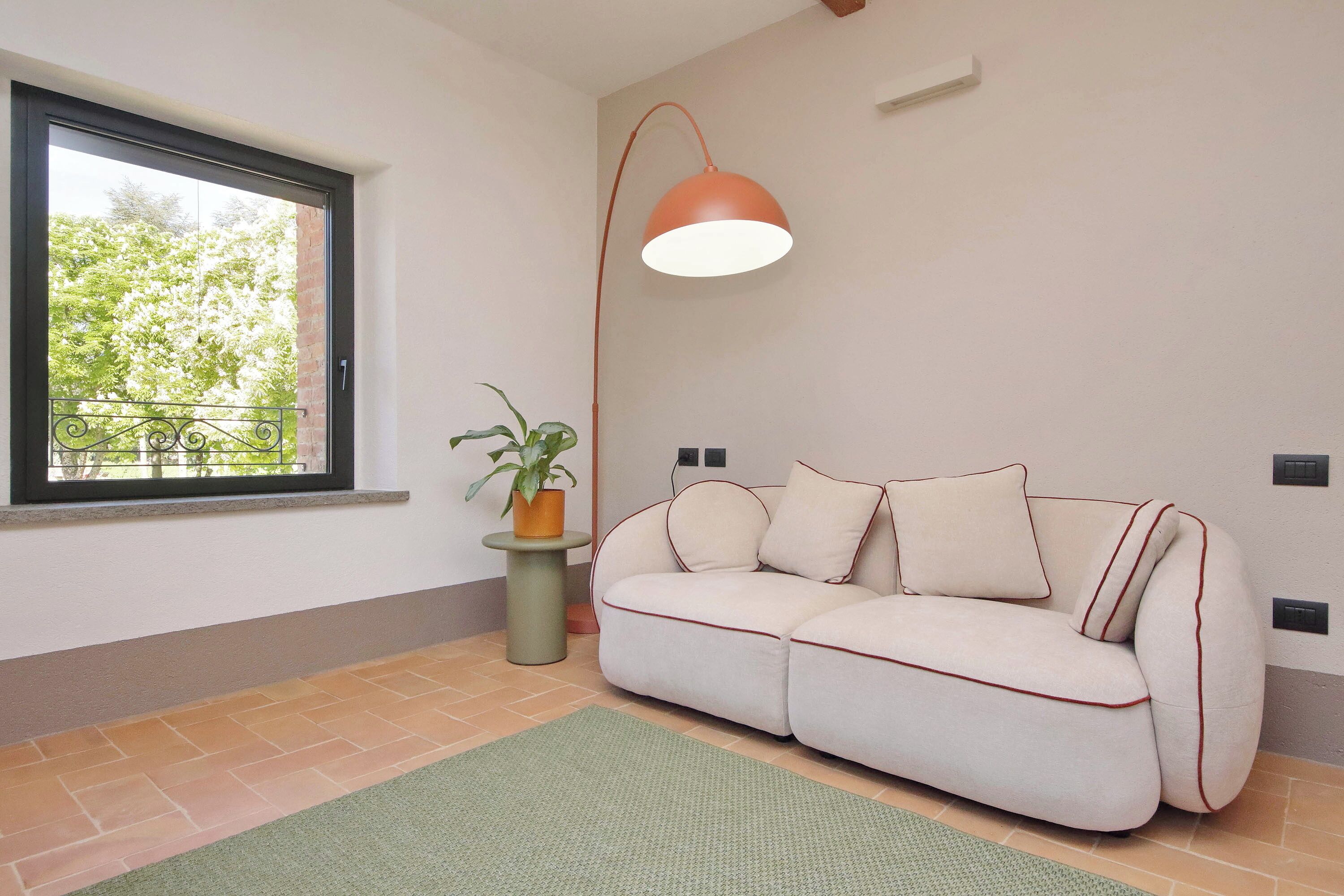 A room with a sofa, a potted plant, a floor lamp, and a window with a view of trees.