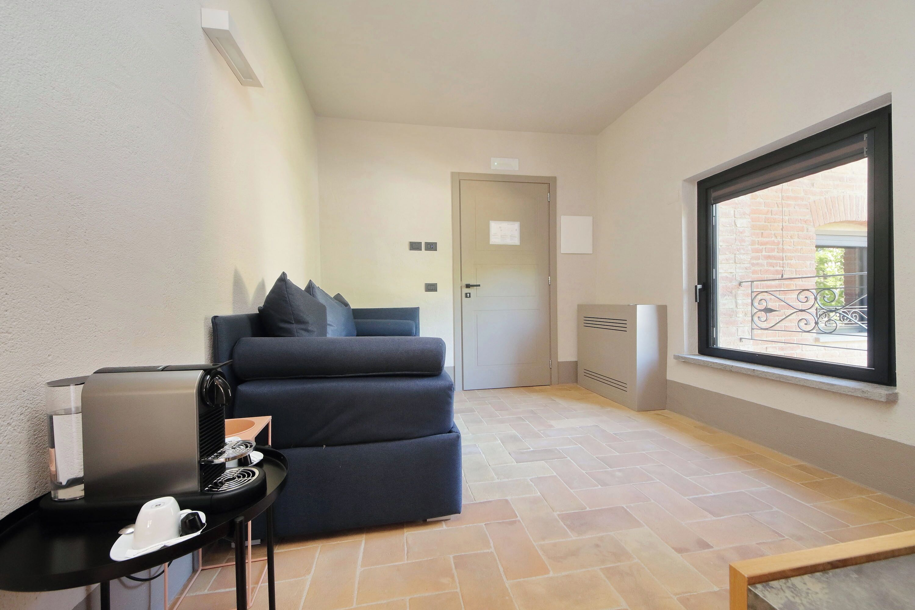 Habitación pequeña con cama, una mesita con cafetera, ventana con vistas y puerta que da a otra habitación.