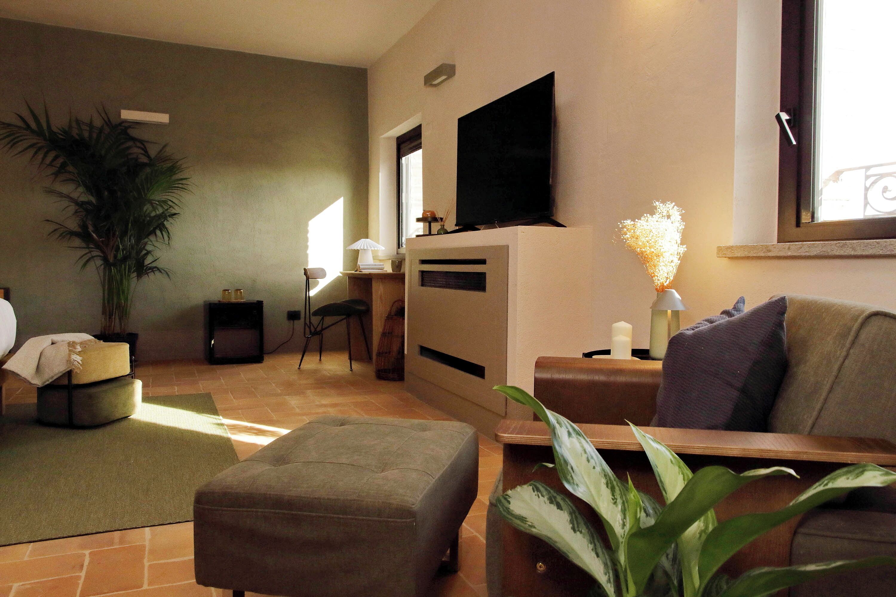 A living room with a sofa, ottoman, a flat-screen TV mounted on the wall, a potted plant, and a window with a view.