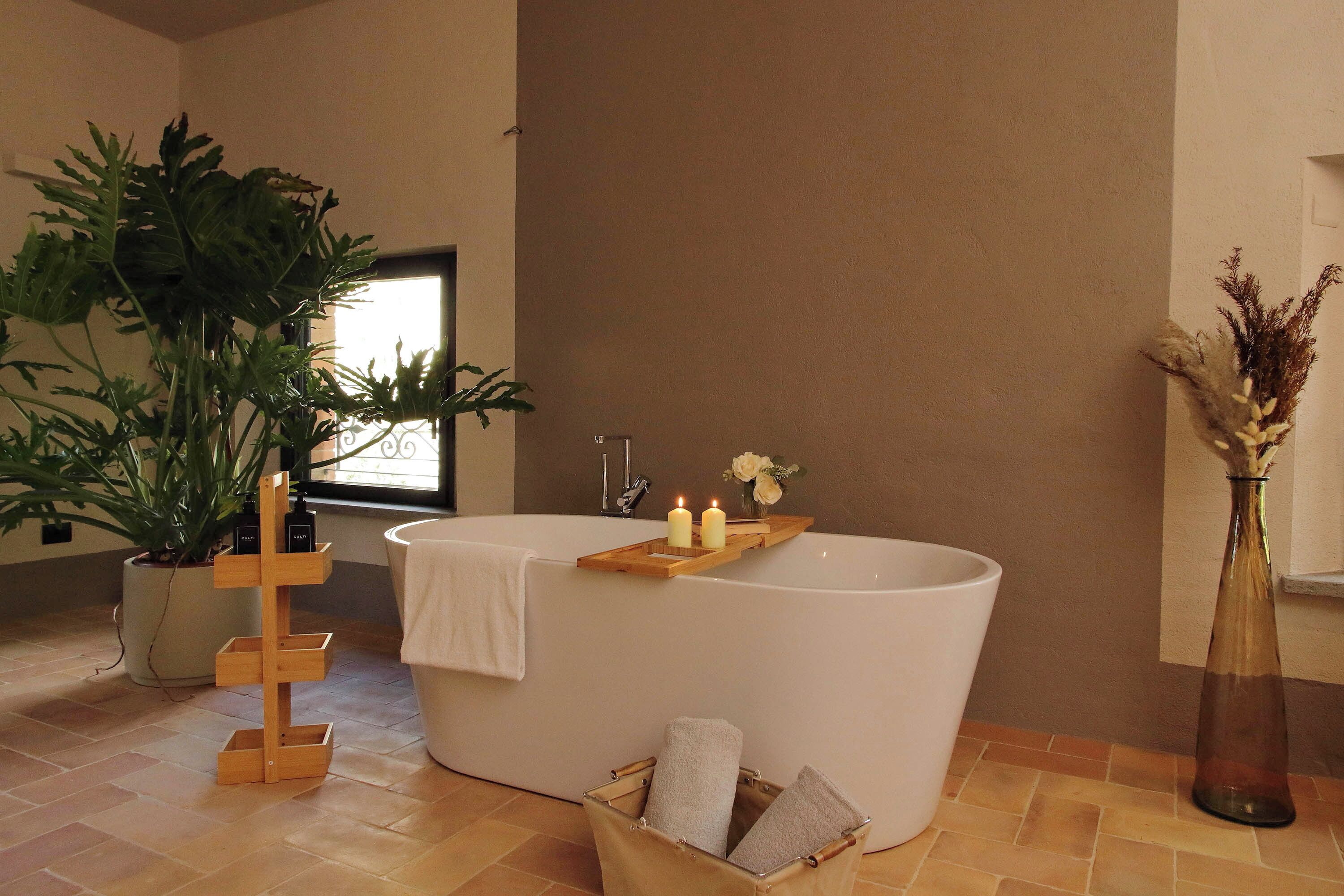 Un bagno moderno con una vasca da bagno indipendente, un supporto per piante in legno, un vaso con fiori secchi e un cesto con asciugamani.