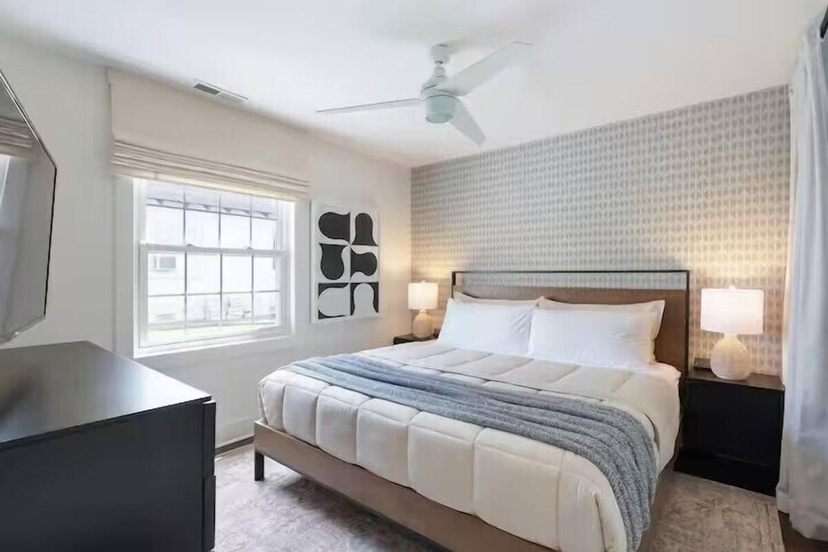 A hotel room with a bed, bedside lamps, a ceiling fan, a window, and a wall with a patterned wallpaper.