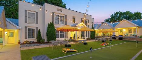 A well-lit outdoor area with string lights, a grassy lawn, and outdoor furniture under umbrellas.
