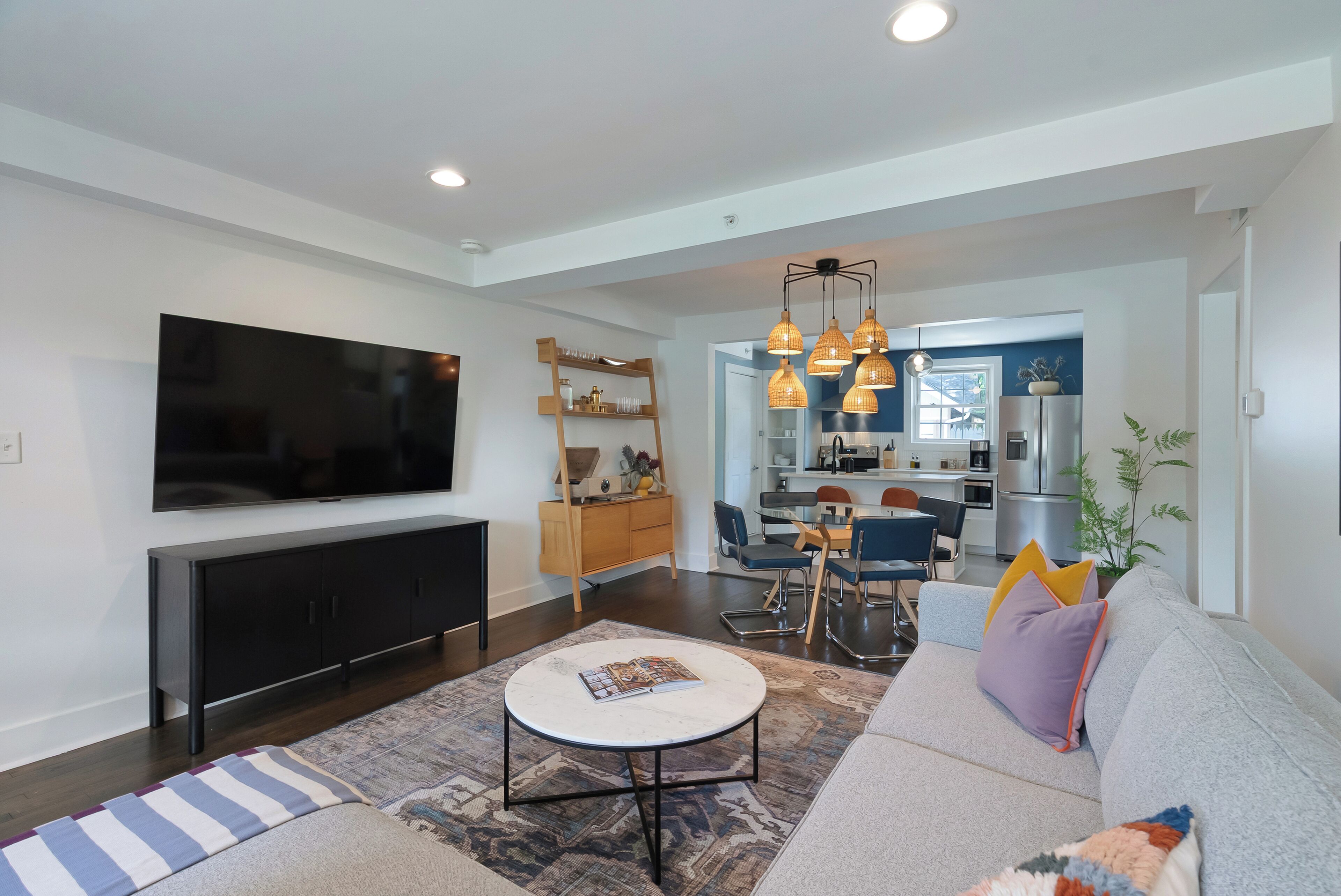 A modern living room with a flat-screen TV, a sofa, a coffee table, and a dining area with chairs and a kitchen in the background.