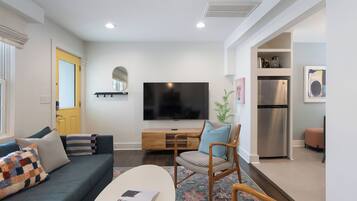 A modern living room with a sofa, a coffee table, a TV, and a kitchen area with a fridge and microwave.