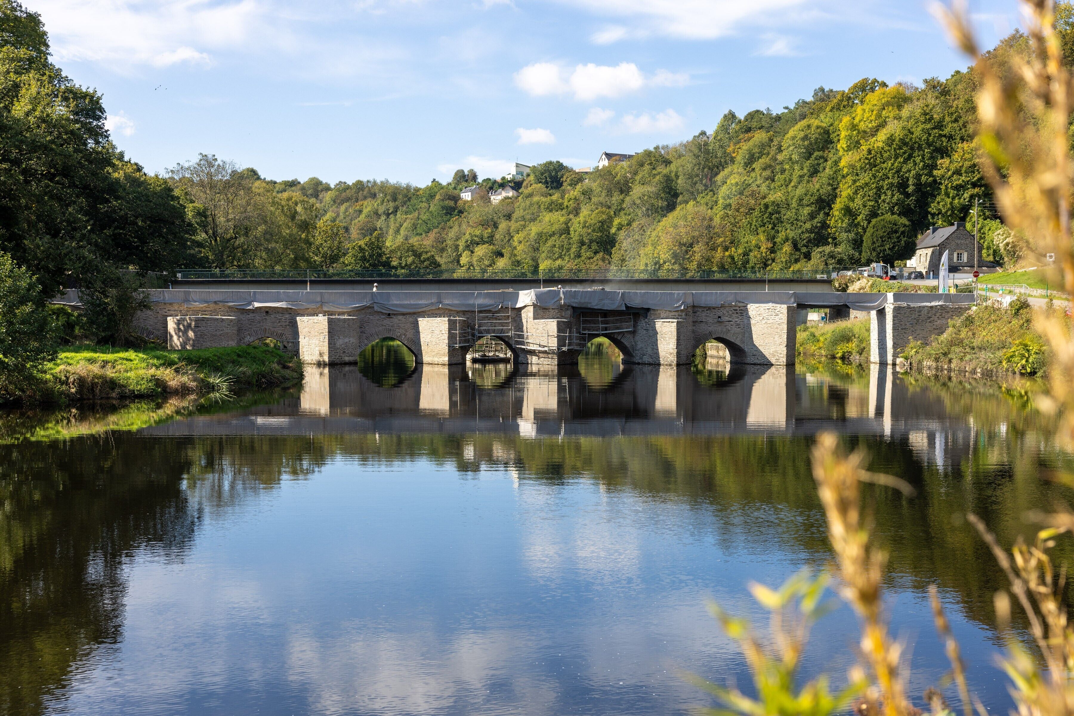 Een stenen brug over een kalme rivier, met een bebost heuvellandschap op de achtergrond.