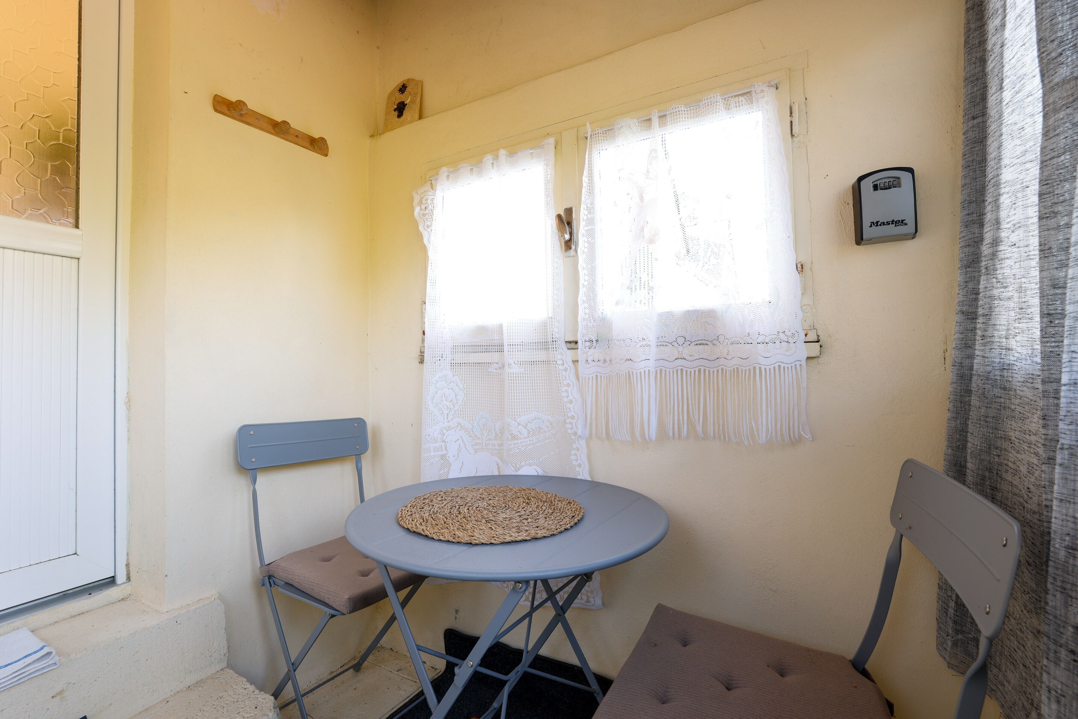 A small room with a round table, two chairs, a window with lace curtains, and a thermostat on the wall.