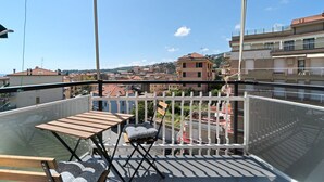Een balkon met een houten tafel en een opklapbare stoel, met uitzicht op een stadsgezicht met gebouwen en een heldere lucht.