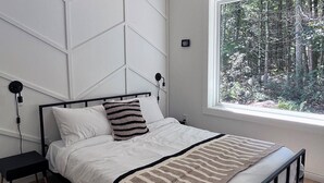 Ein modernes Schlafzimmer mit Bett, Deckenventilator, Fenster mit Blick auf einen Wald und einem Nachttisch.