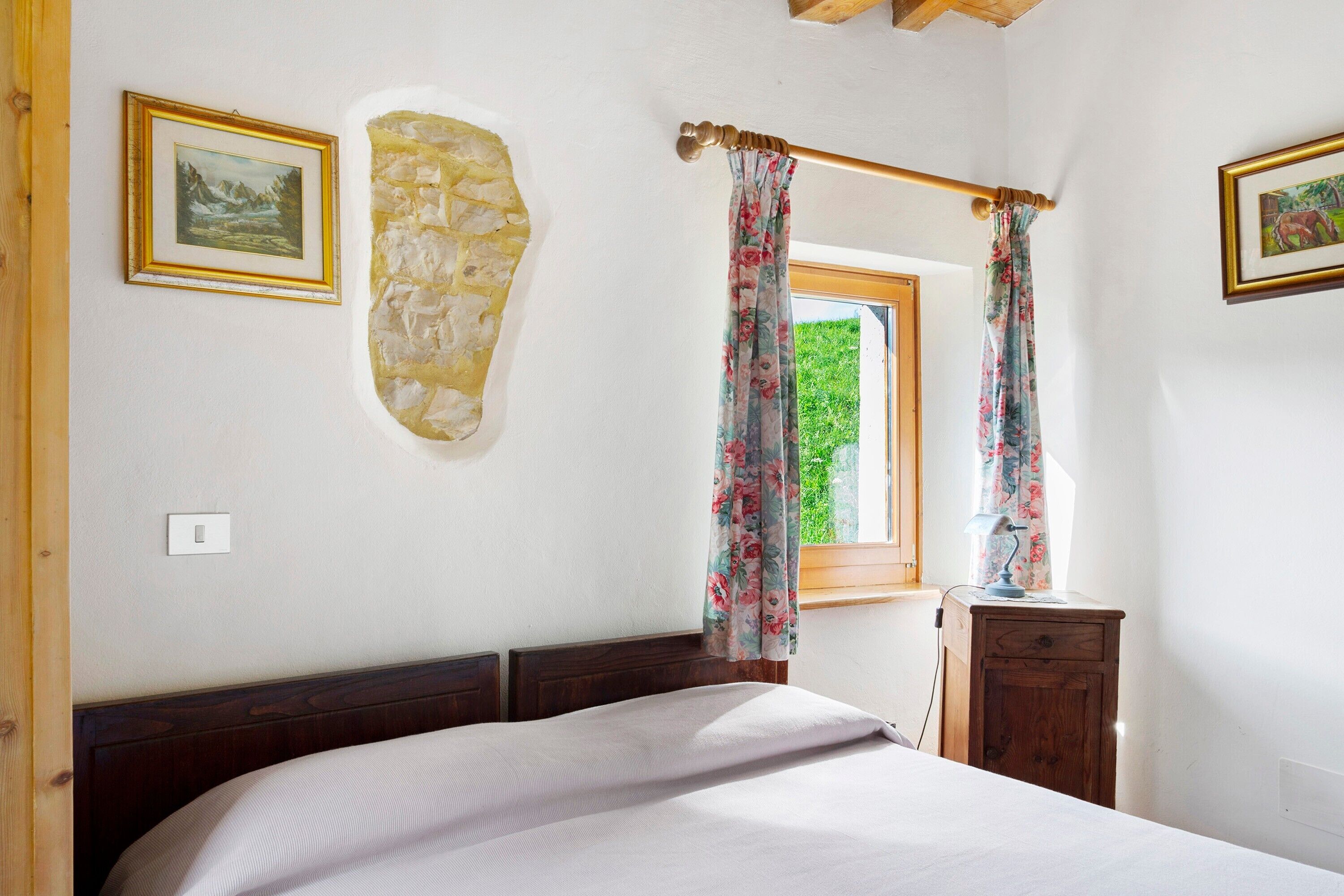 A bedroom with a bed, a wooden headboard, a stone wall, a window with curtains, and a small wooden nightstand.