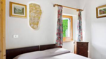 A bedroom with a bed, a wooden headboard, a stone wall, a window with curtains, and a small wooden nightstand.