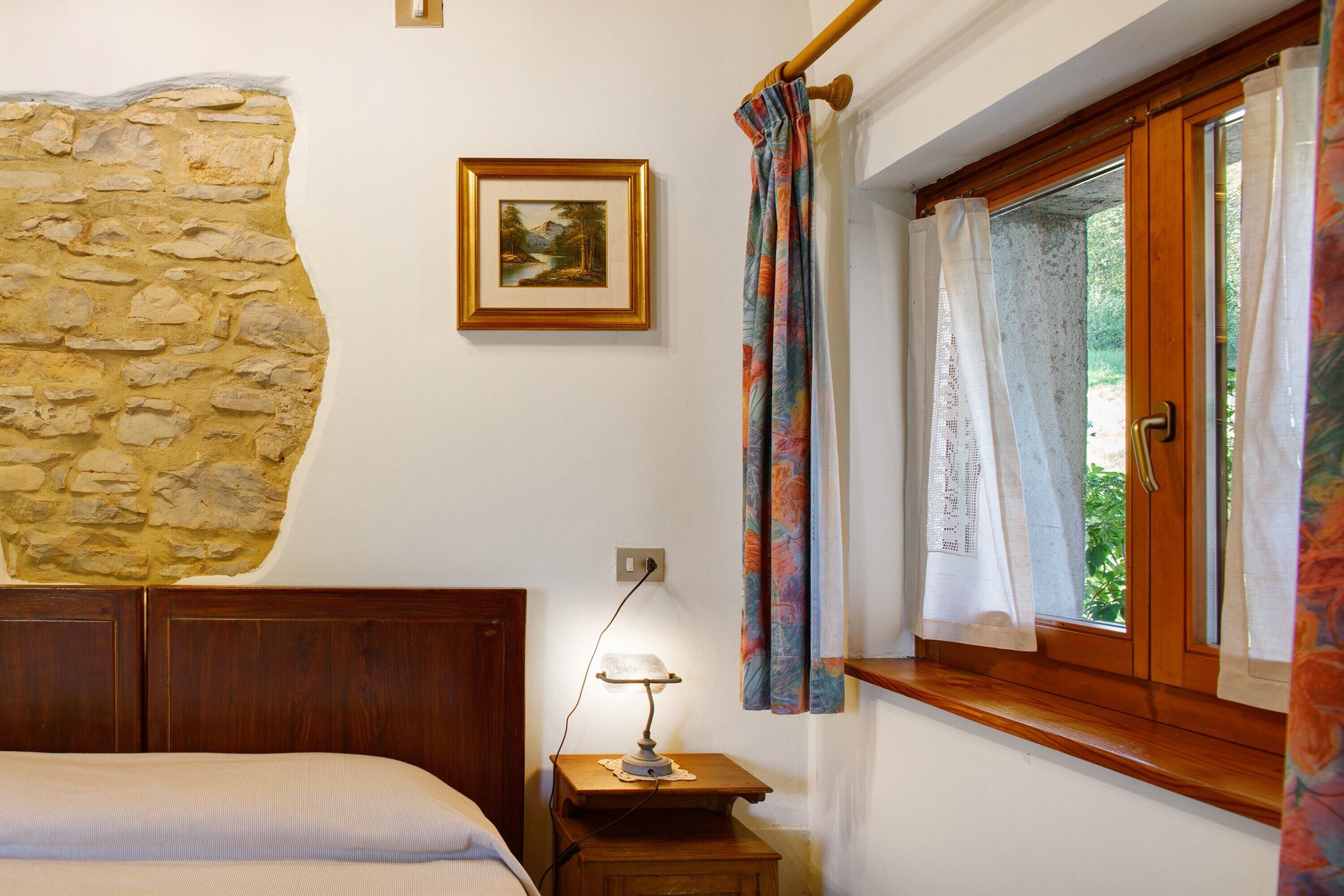 Quarto com parede de pedra, cabeceira de madeira, mesa de cabeceira com abajur, janela com cortinas e um quadro na parede.